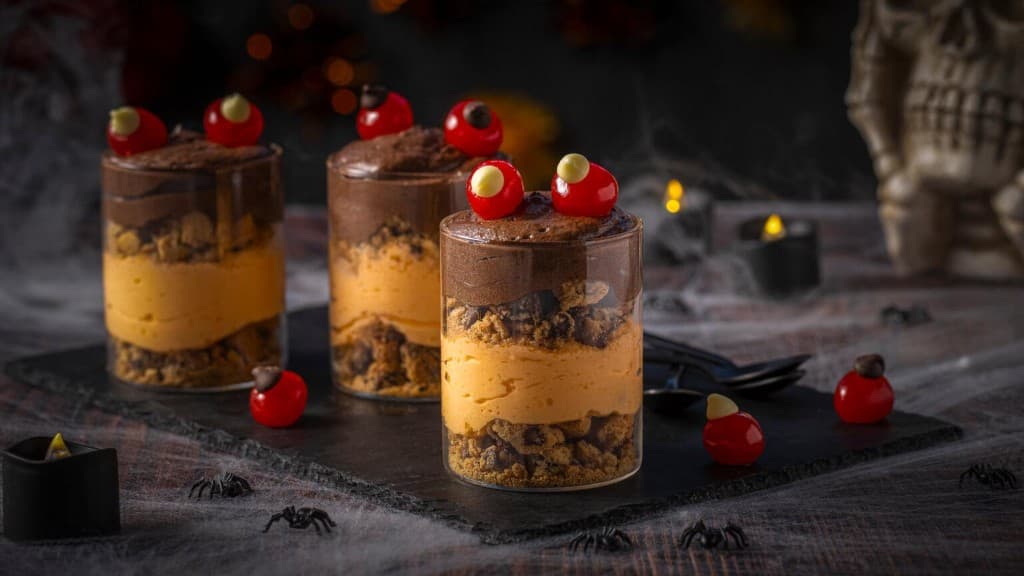 Three glasses of layered cacao mousse decorated with candy eyes on a dark Halloween table