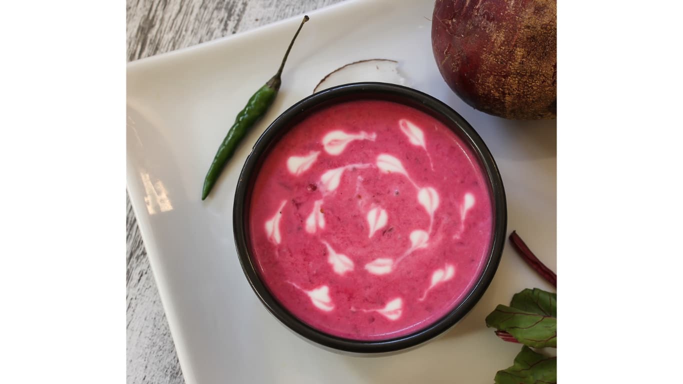 A black bowl of bright pink beet soup with a white coconut-milk swirl, next to a whole beet, beet greens, and a green chili