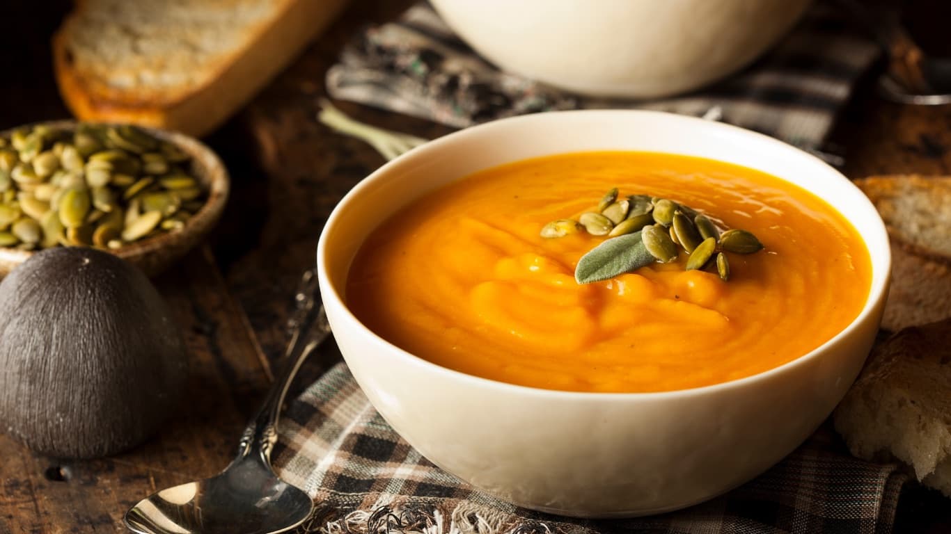 A bowl of orange squash soup topped with pumpkin seeds and a sage leaf, with bread and a bowl of toasted seeds on a wooden table