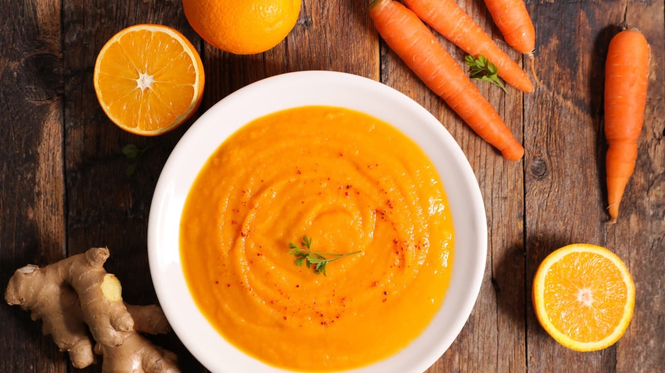 A white bowl of bright orange carrot soup with a parsley sprig, surrounded by carrots, ginger, and orange halves on a wooden table