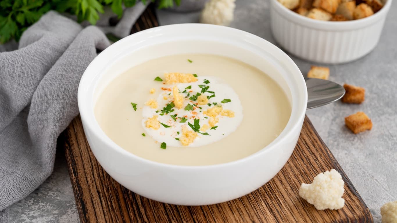 A white bowl of pale cauliflower soup with a coconut-milk swirl and herbs, with cauliflower florets and croutons nearby