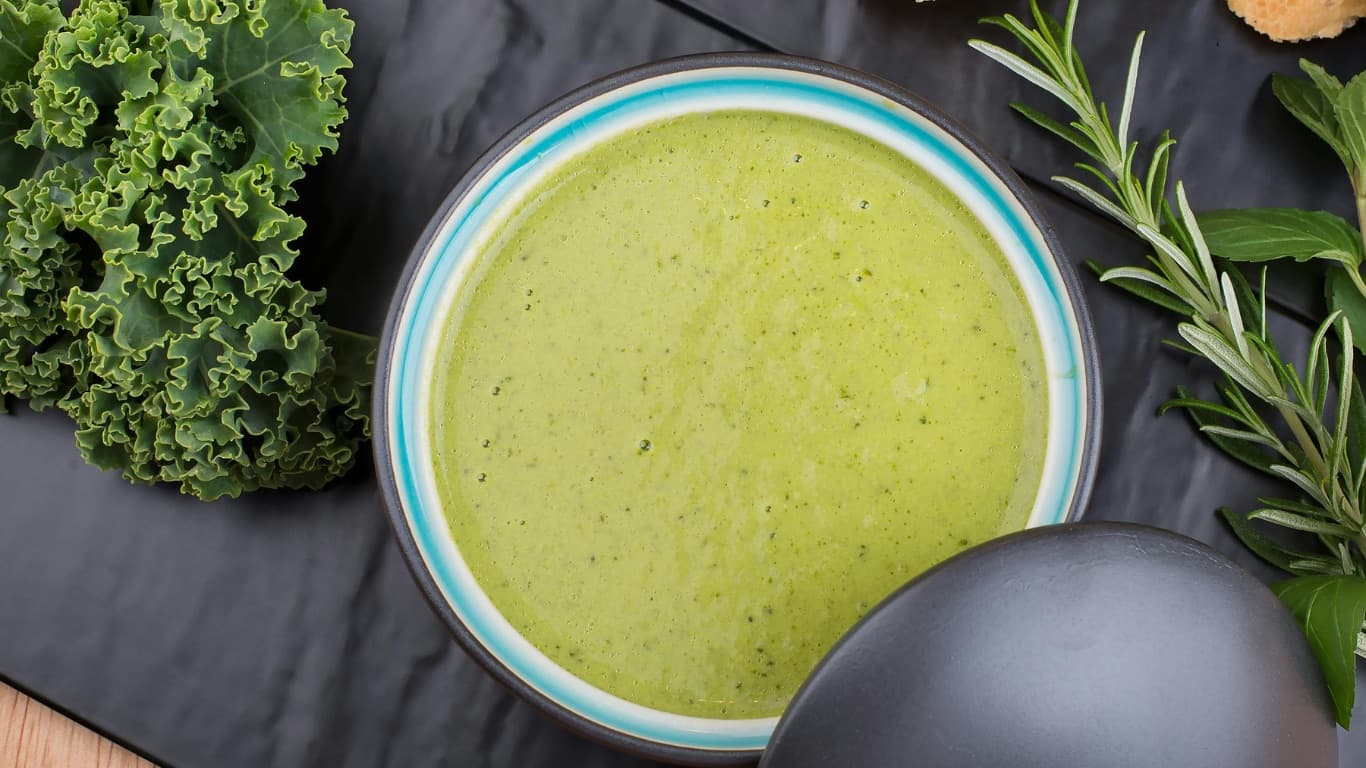 A bowl of pale green kale soup beside curly kale leaves and sprigs of rosemary