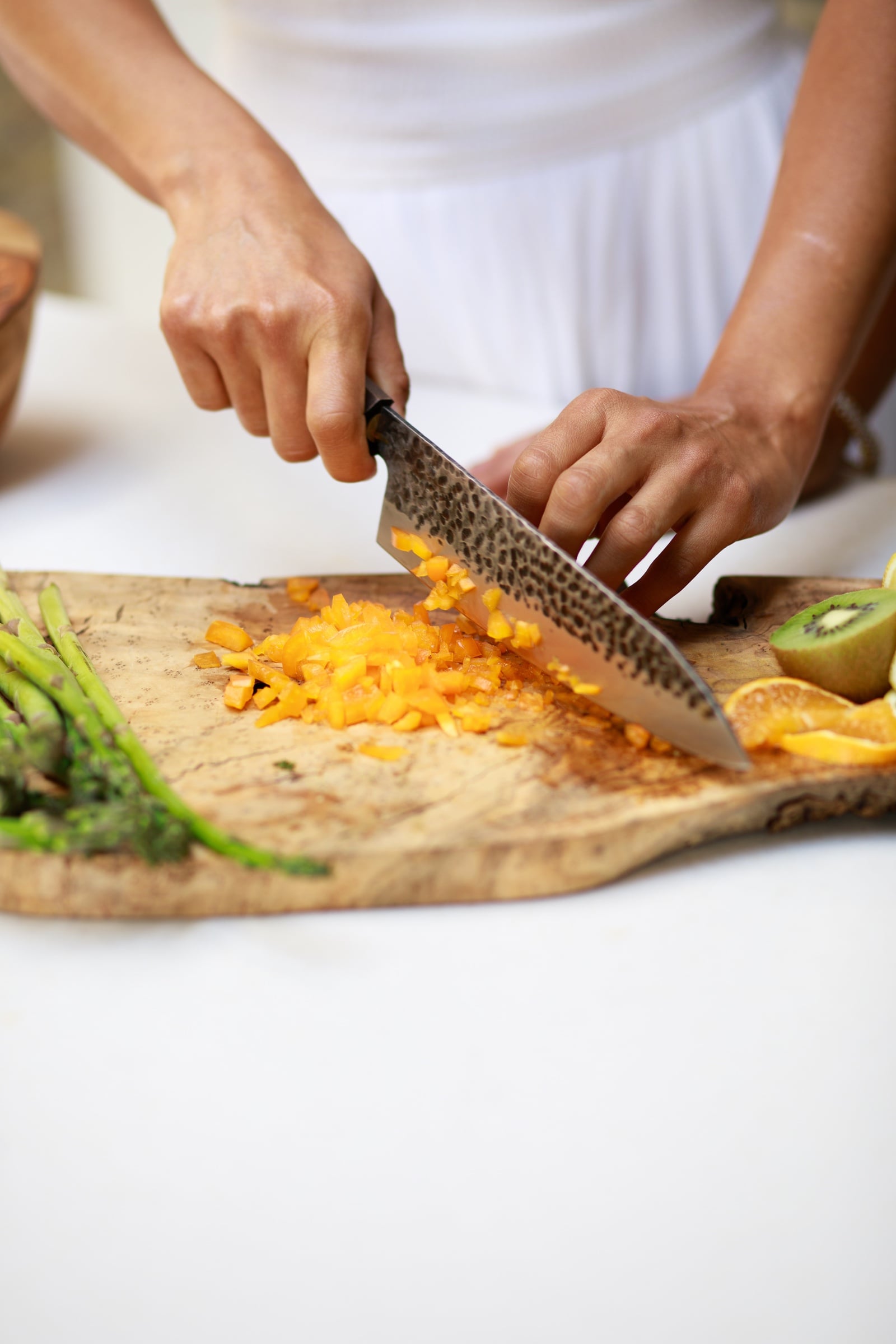 Hands chopping orange vegetables on a wooden cutting board with asparagus, kiwi, and citrus