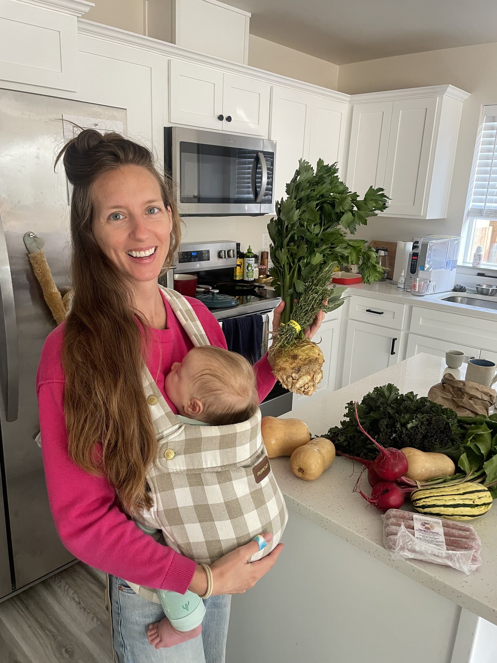 Madelaine Eschrich in her kitchen with a baby in a carrier, holding fresh celeriac